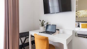 A hotel room with a white desk, a laptop, a small potted plant, and a bed with a patterned headboard.