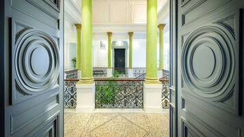 A grand hallway with high green columns, a decorative iron railing, and a patterned floor.