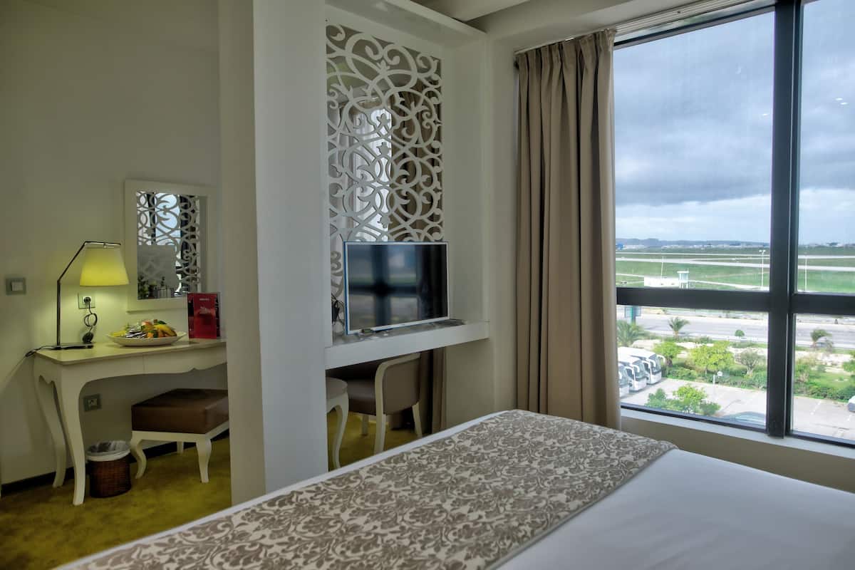 A hotel room with a bed, a desk with a TV, a lamp, and a view of an airport.