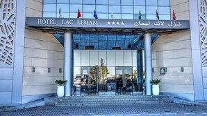The entrance of the HOTEL LAC LEMAN with a modern facade, large glass doors, and multiple flags displayed above. - Hôtel Lac Léman, LES BERGES DU LAC (Tunis)