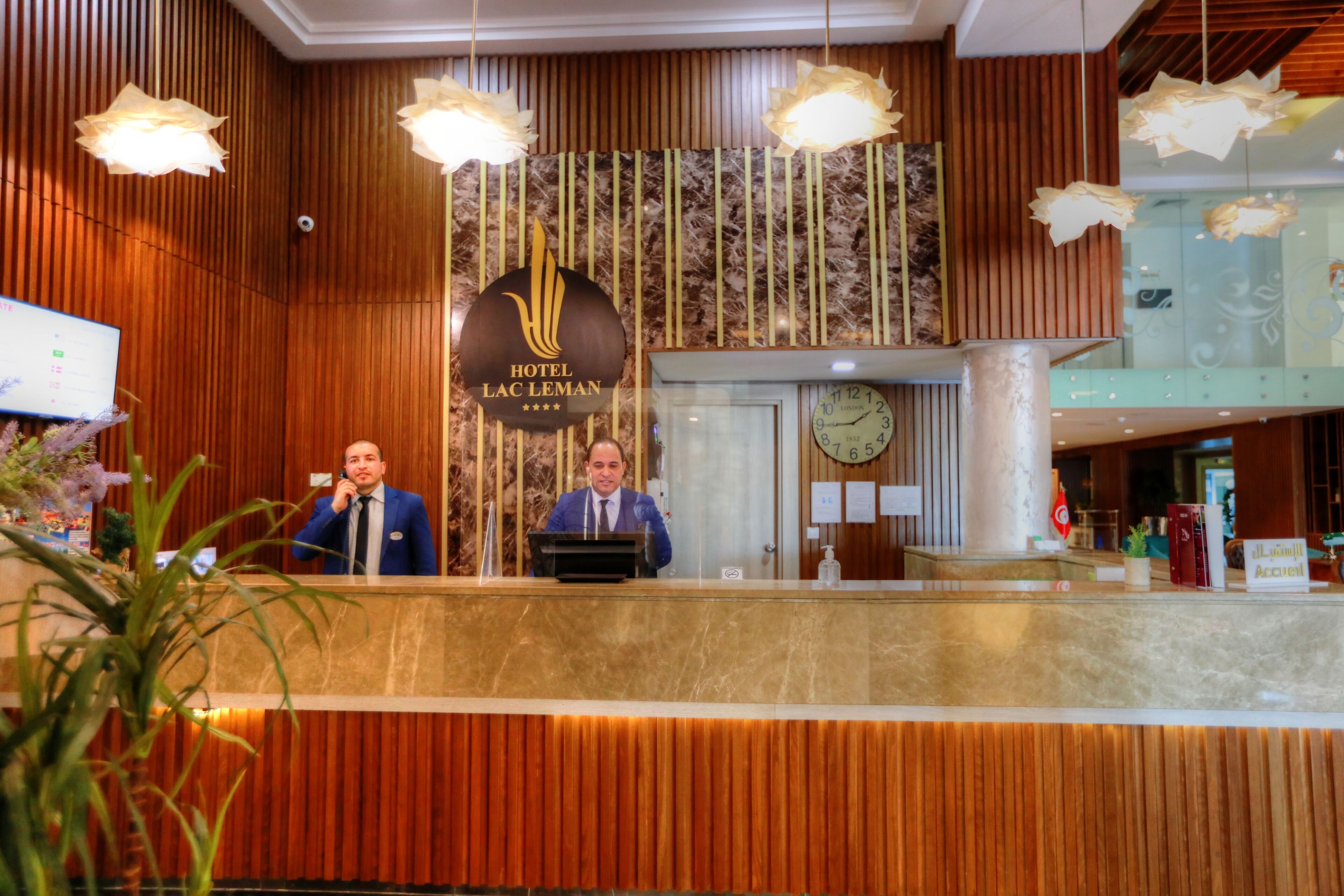 A hotel reception desk with a wooden panel design, a clock, and a sign indicating the hotel name.