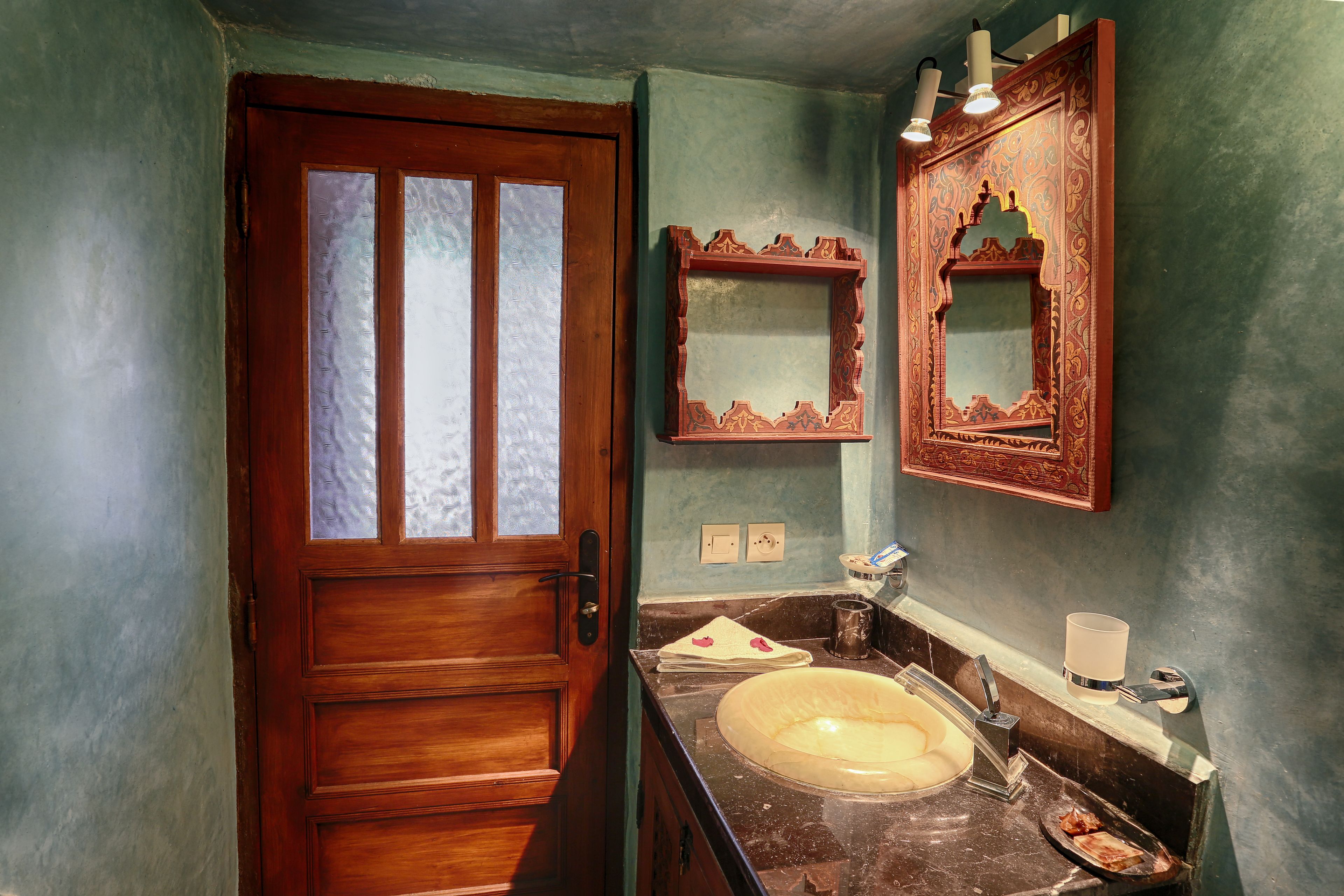 Un baño con puerta de madera, un lavabo, un espejo y una lámpara fijada a la pared.