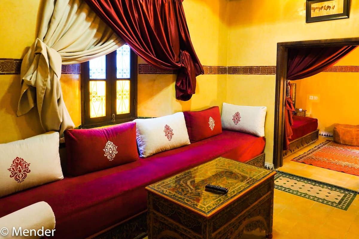 Una habitación tradicional marroquí con un sofá rojo, cojines estampados, una mesa de centro de madera y un espejo decorativo.