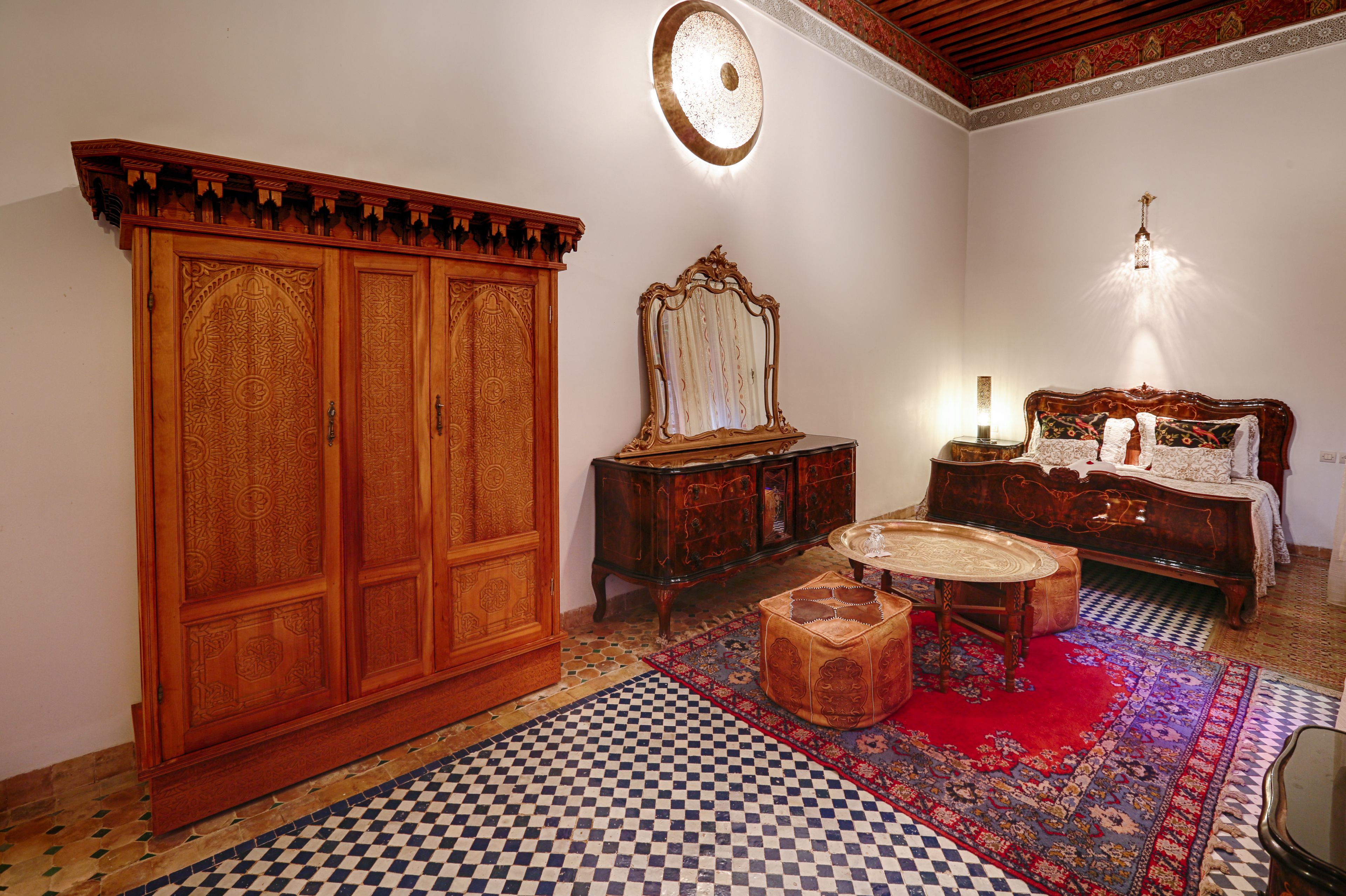 A traditional room with a large wooden wardrobe, a bed with ornate headboard, a round wooden table, and a patterned rug.