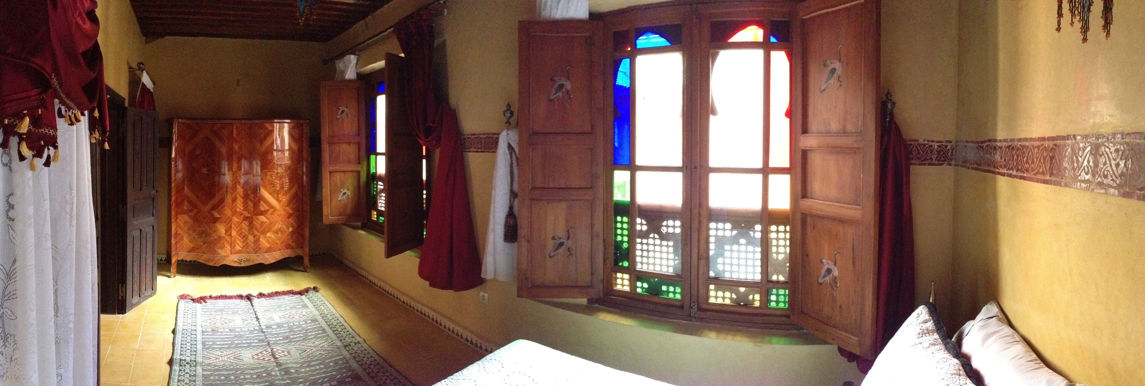 A room with a patterned rug, wooden wardrobe, and a window with stained glass.