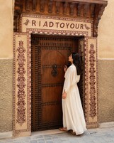 Una mujer de pie frente a una puerta de madera con la inscripción 'RIAD TOYOUUR' y una decoración ornamental.