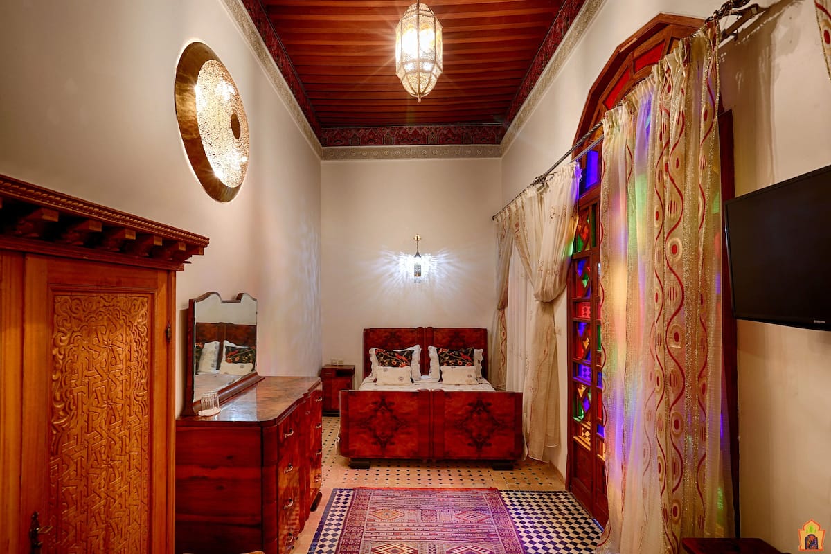 Un dormitorio con armario de madera, cama con ropa de cama estampada, espejo y una araña.