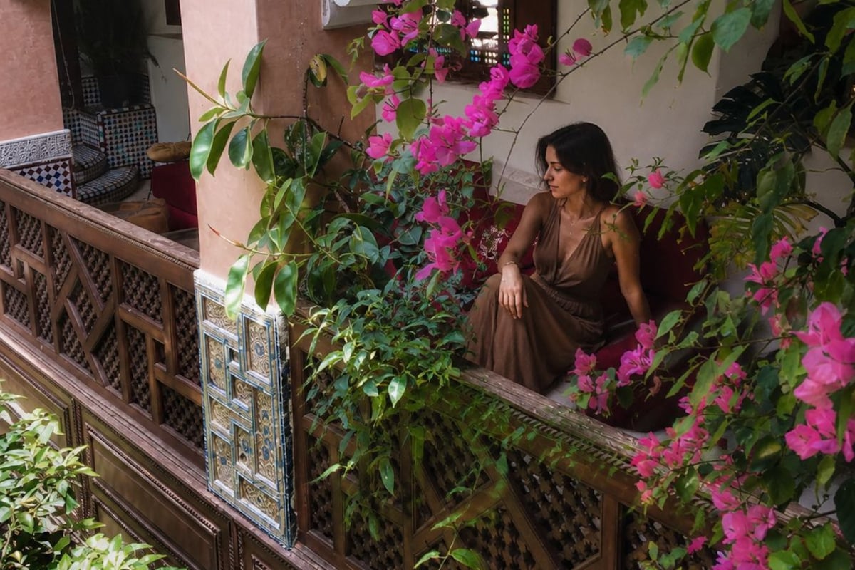 Un balcón con una persona sentada, rodeado de exuberante follaje y flores rosadas.