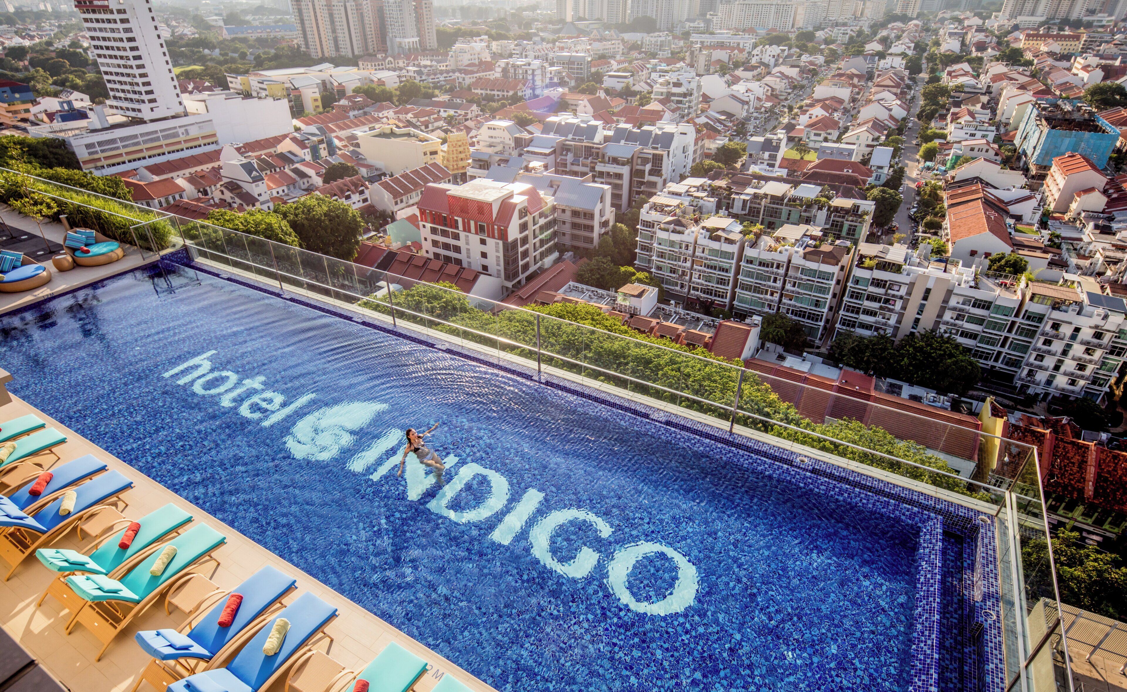 A rooftop pool with 'hotel INDIGO' written on the water, surrounded by lounge chairs and a cityscape view.
