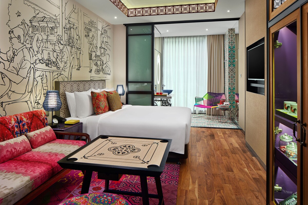A hotel room with a bed, a sofa, a table, a TV, and a bathroom with a mirror and shelves.