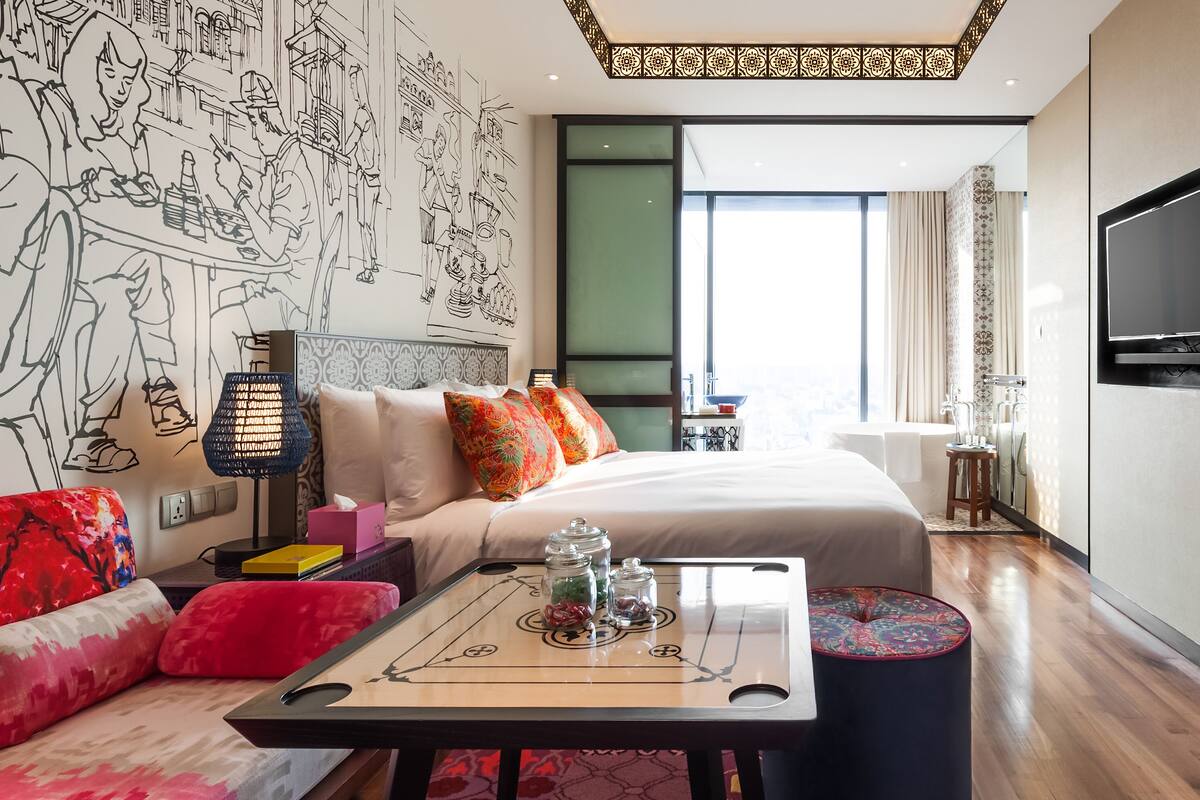 A modern hotel room with a large bed, a desk with a board game, a TV, and a mural on the wall.