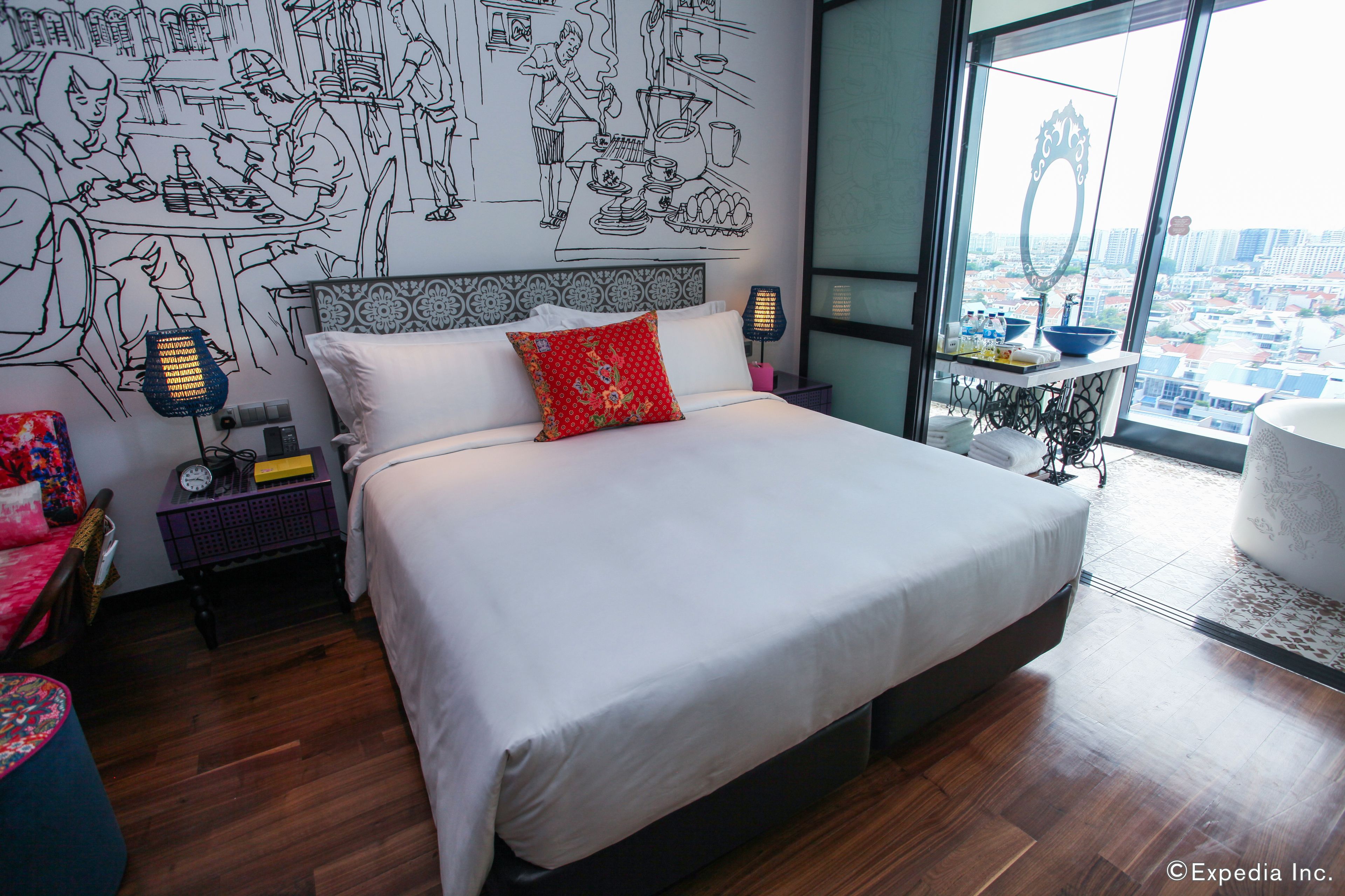 Una habitación de hotel moderna con una cama grande, vistas a la ciudad, una zona de comedor con un mural en blanco y negro, y una pequeña cocina.