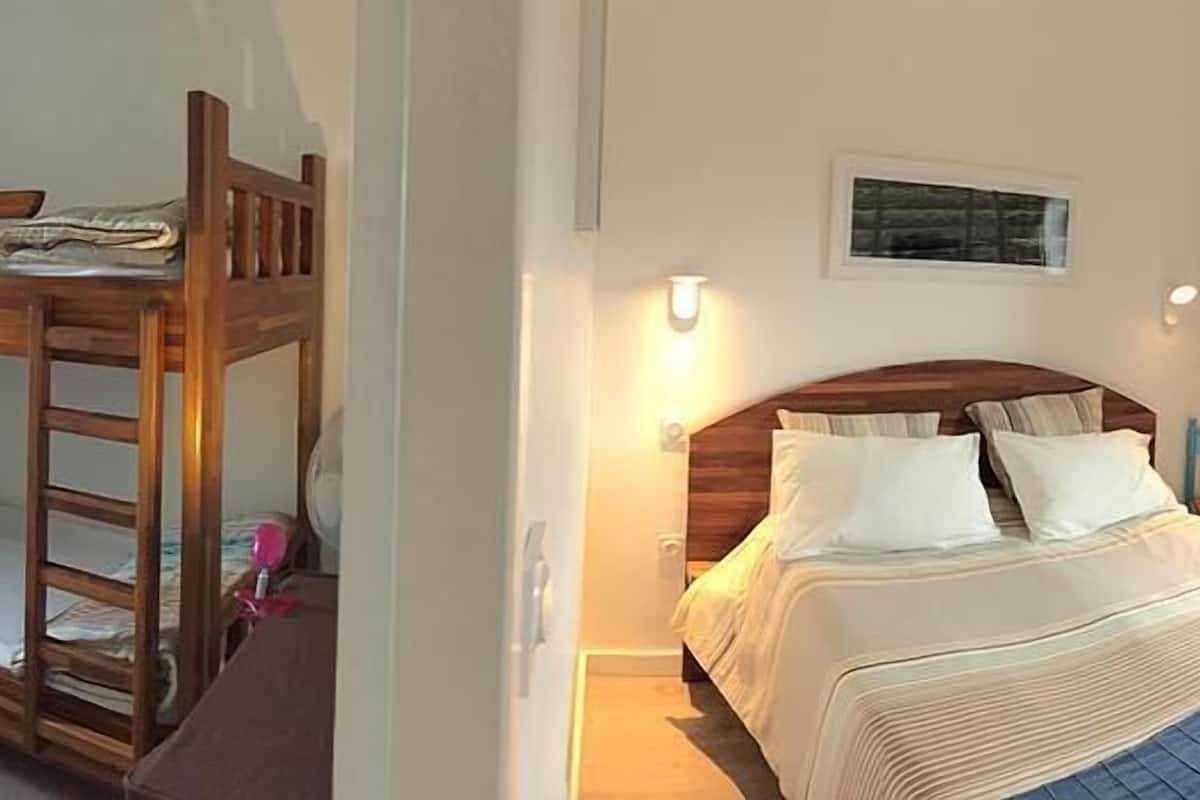 Une chambre avec un lit superposé et un lit simple, tous deux dotés de draps blancs. Un luminaire mural est fixé au-dessus du lit simple, et un tableau encadré orne le mur.
