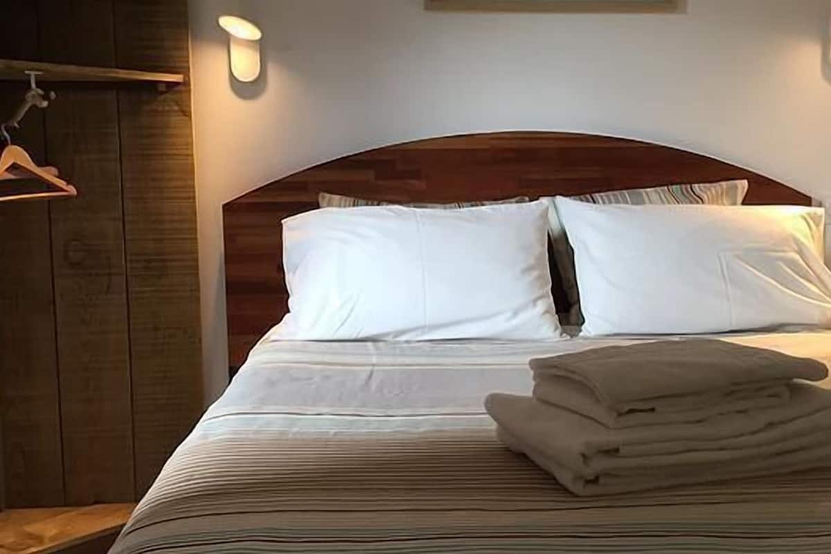 Un lit bien fait, avec du linge de lit blanc et une taie d’oreiller à rayures, une tête de lit en bois et un luminaire fixé au mur.