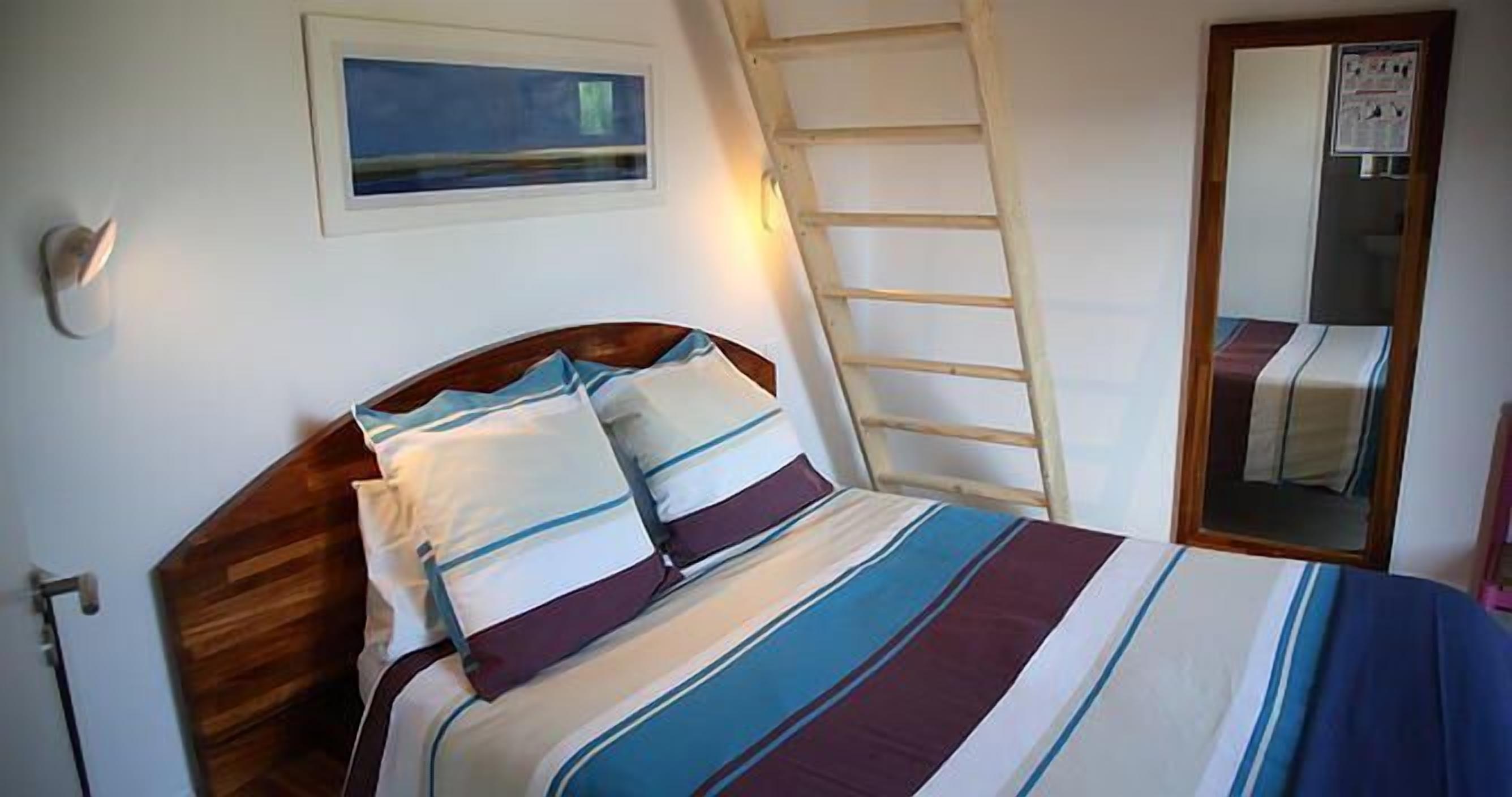 Un dormitorio con cabecera de madera, una cama con ropa de cama a rayas, una escalera apoyada en la pared y un cuadro enmarcado sobre la cama.