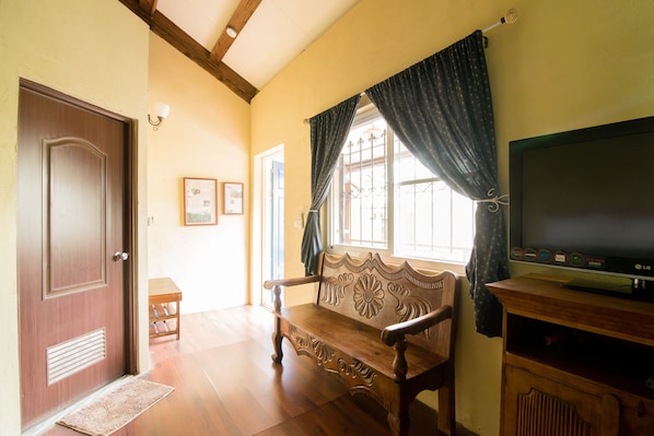 Una habitación con un banco de madera, un televisor sobre un mueble de madera, una puerta y una ventana con cortinas.