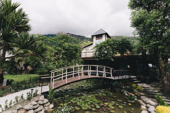 A wooden bridge over a pond with lily pads, surrounded by lush greenery and a building with a peaked roof.