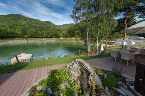 A lakeside deck with outdoor seating, a small waterfall feature, and a view of the lake and surrounding hills.