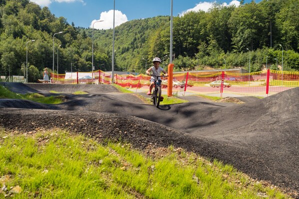 A motocross track with a rider in the center, surrounded by orange safety netting and a forested hillside in the background.