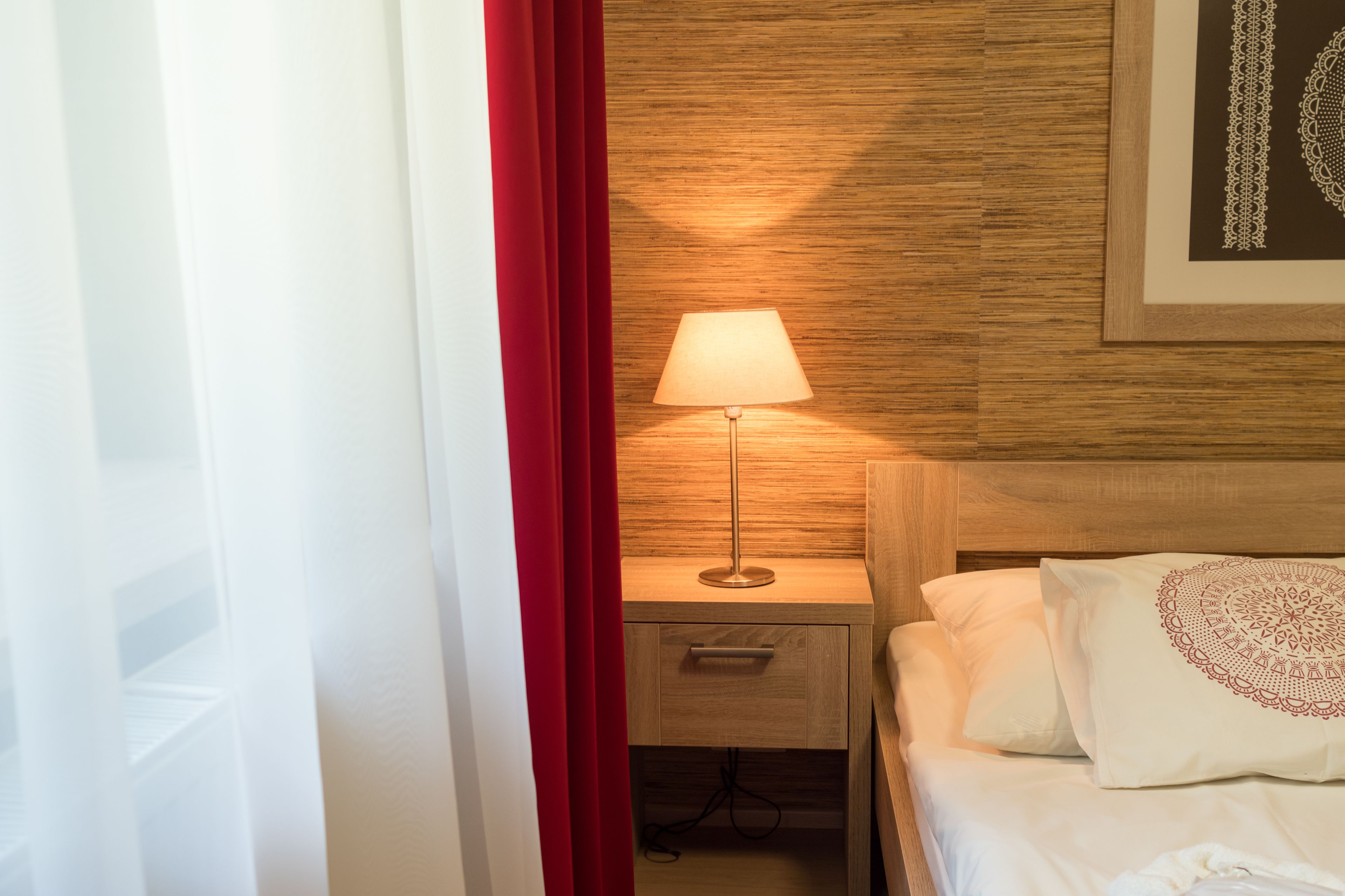 A hotel room with a wooden headboard, a bedside table with a lamp, a drawer, and a bed with pillows.