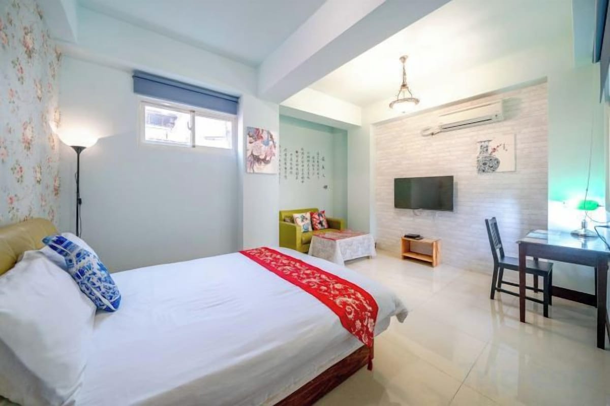 A bedroom with a bed, a desk, a chair, a television, and a window.