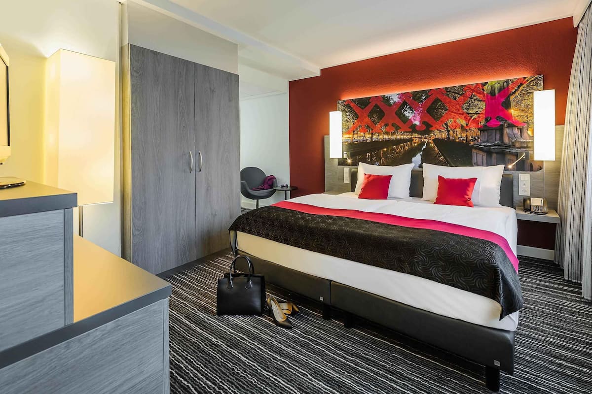 Quarto de hotel com cama, mesas de cabeceira, escrivaninha, guarda-roupa e parede com padrão decorativo.