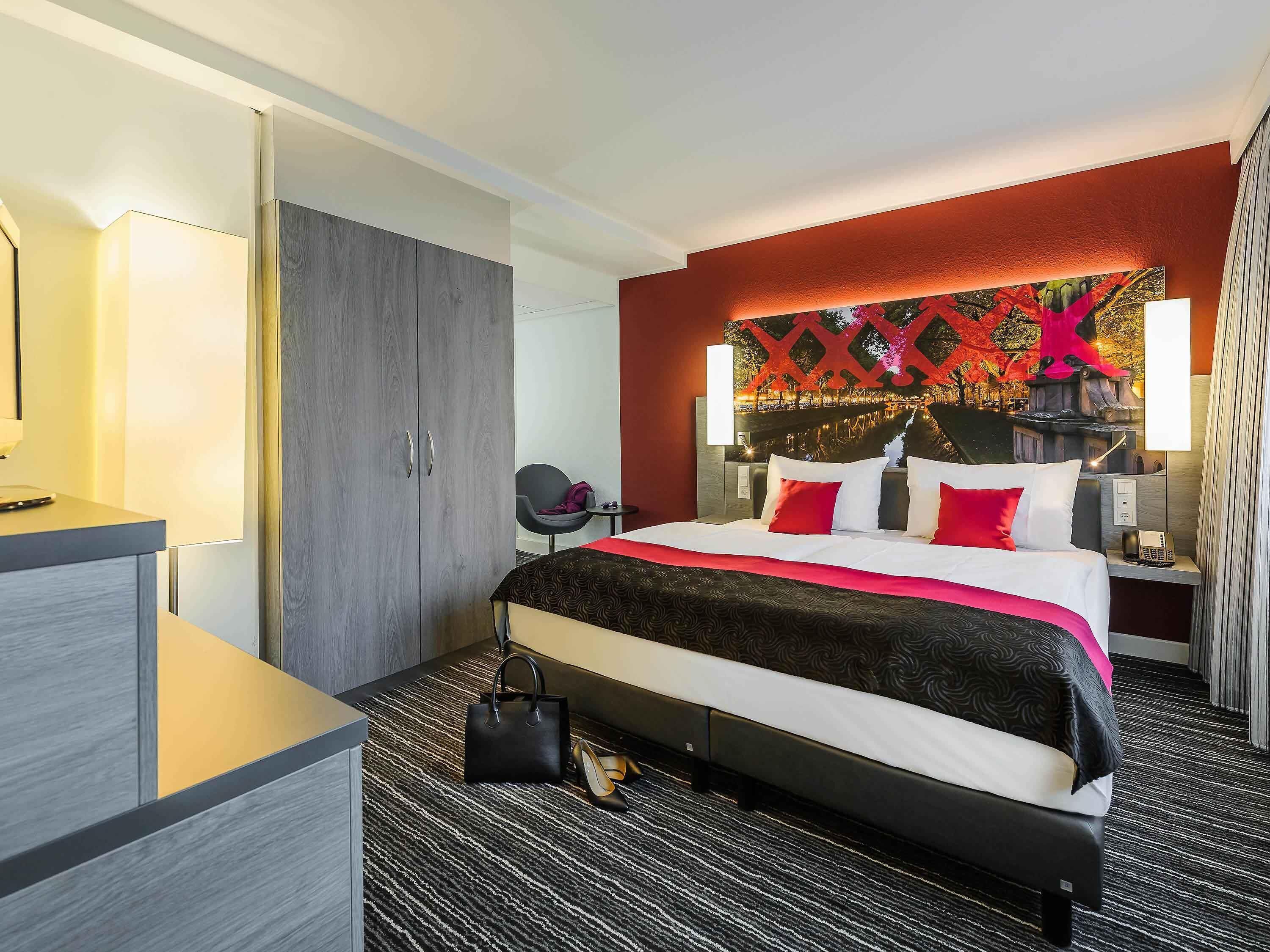 A hotel room with a bed, bedside table, lamp, and a view of a cityscape.