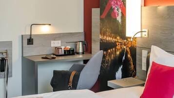 Ein modernes Hotelzimmer mit einem Bett, einem Schreibtisch mit Wasserkocher, einer Kaffeemaschine und einem großen Fenster mit Blick auf die Stadt.