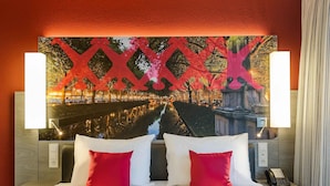 A hotel room with a large bed, two red pillows, a nightstand with a phone, and a wall-mounted TV displaying a cityscape.