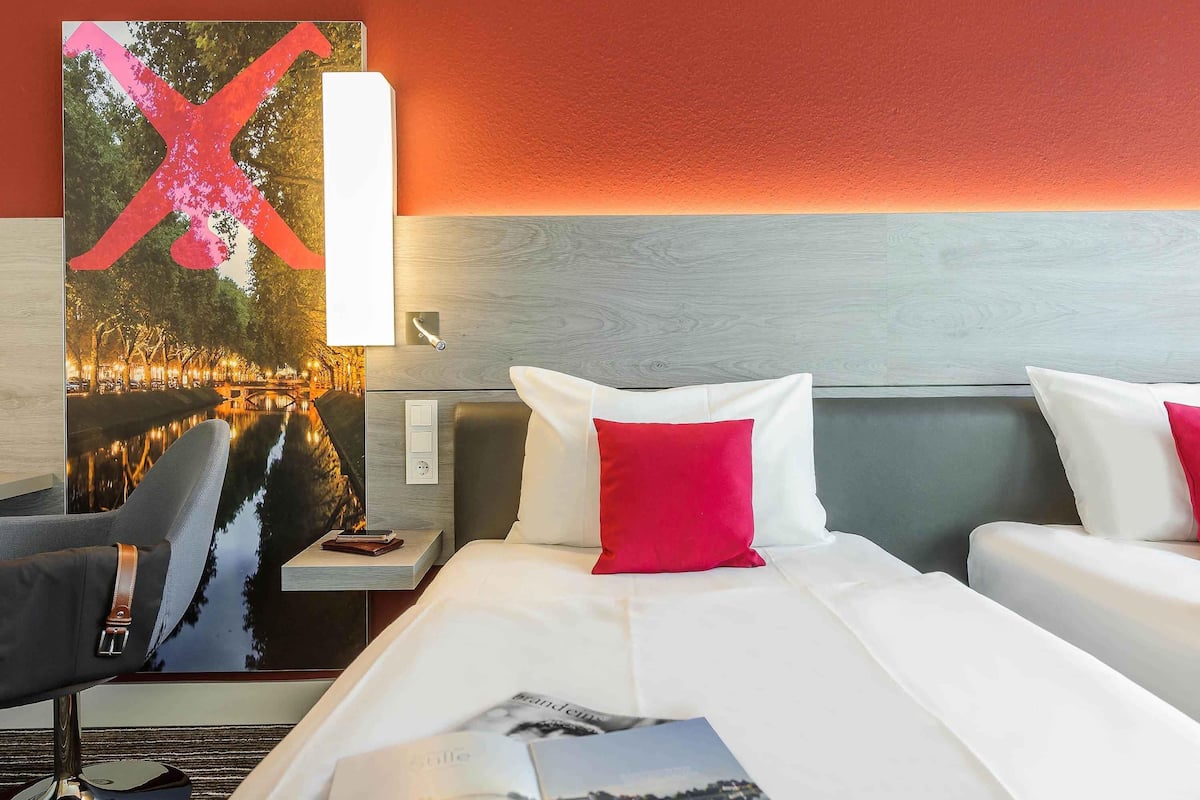 Una habitación de hotel con cama, dos mesitas de noche, una silla, un mueble auxiliar y televisor instalado en la pared.