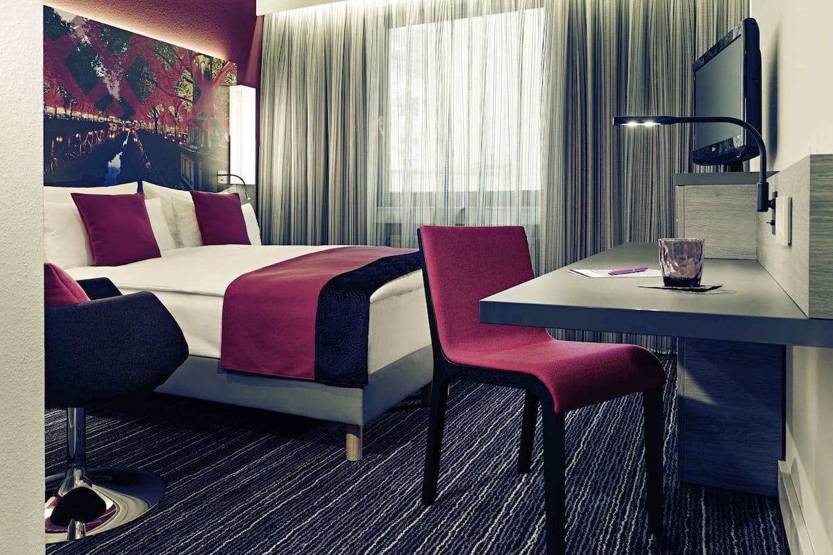 Quarto de hotel com uma cama, uma escrivaninha, uma cadeira, um abajur e uma janela grande com cortinas.
