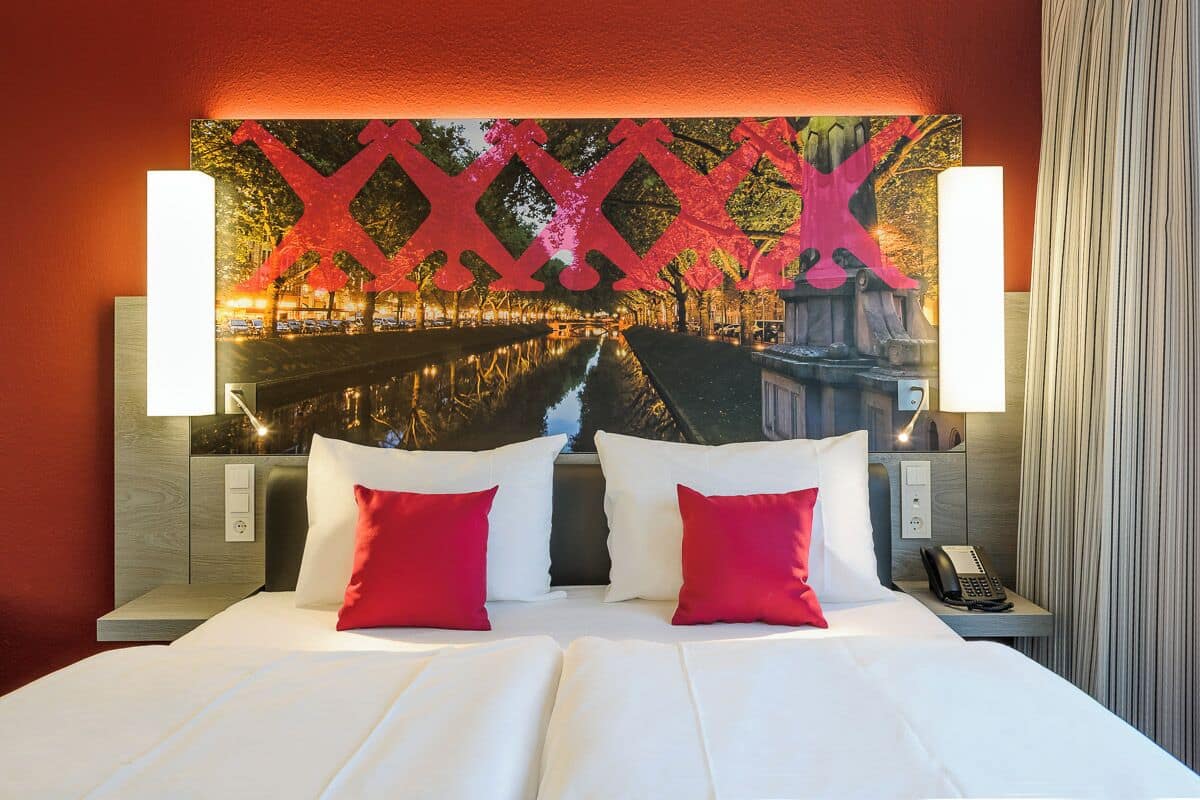 Quarto de hotel com uma cama grande, duas almofadas vermelhas, uma mesa de cabeceira com telefone e uma obra de arte na parede retratando um horizonte urbano.