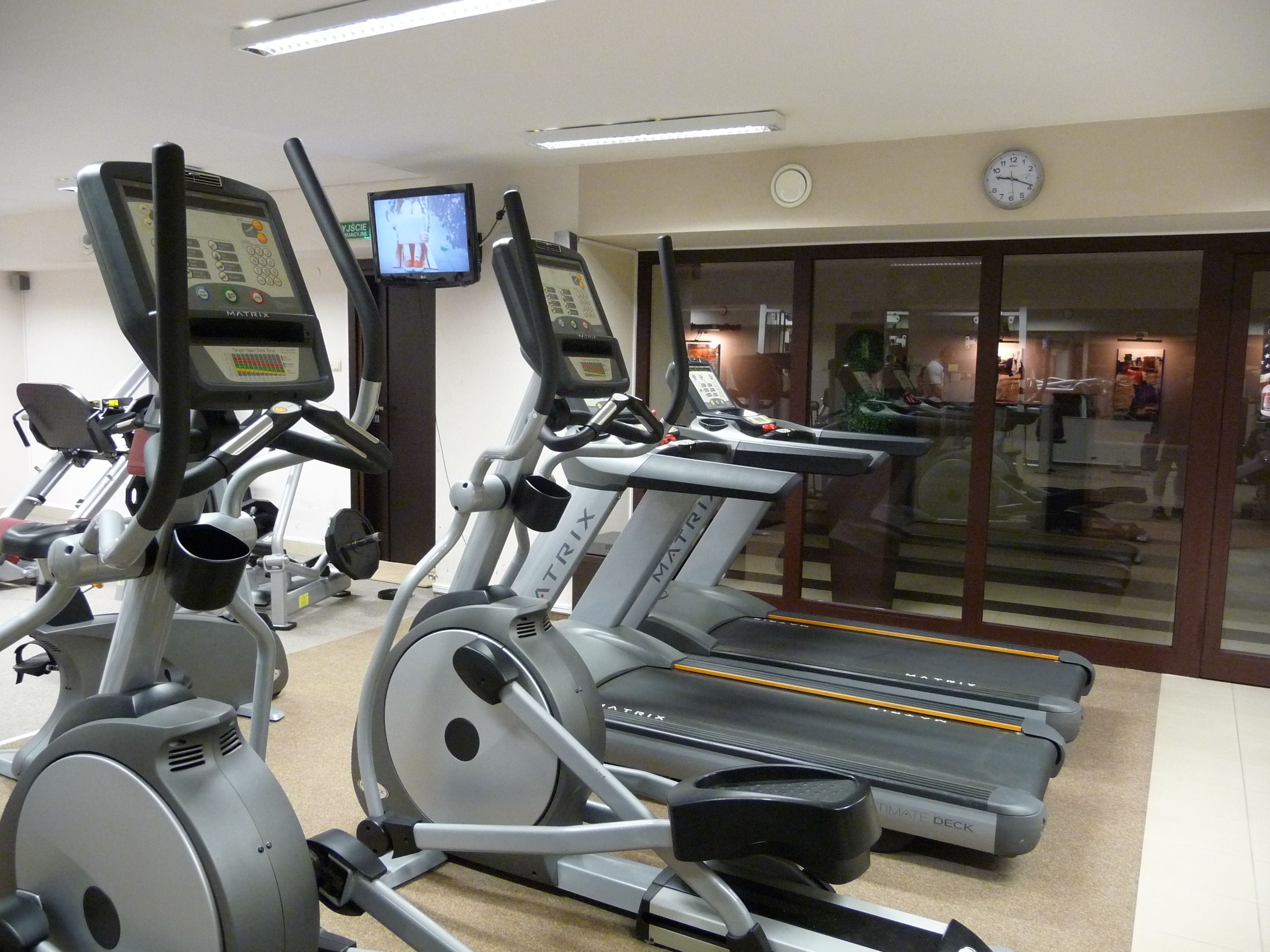 A gym with multiple treadmills, stationary bikes, and a television screen displaying an exercise video.