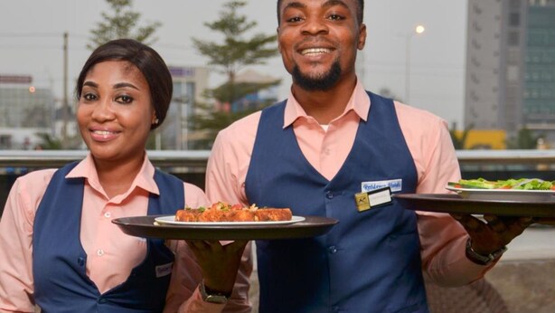 Two waitstaff holding trays of food in an outdoor dining area with a view of buildings in the background. - Residency Hotel Lekki Lagos (Lekki)
