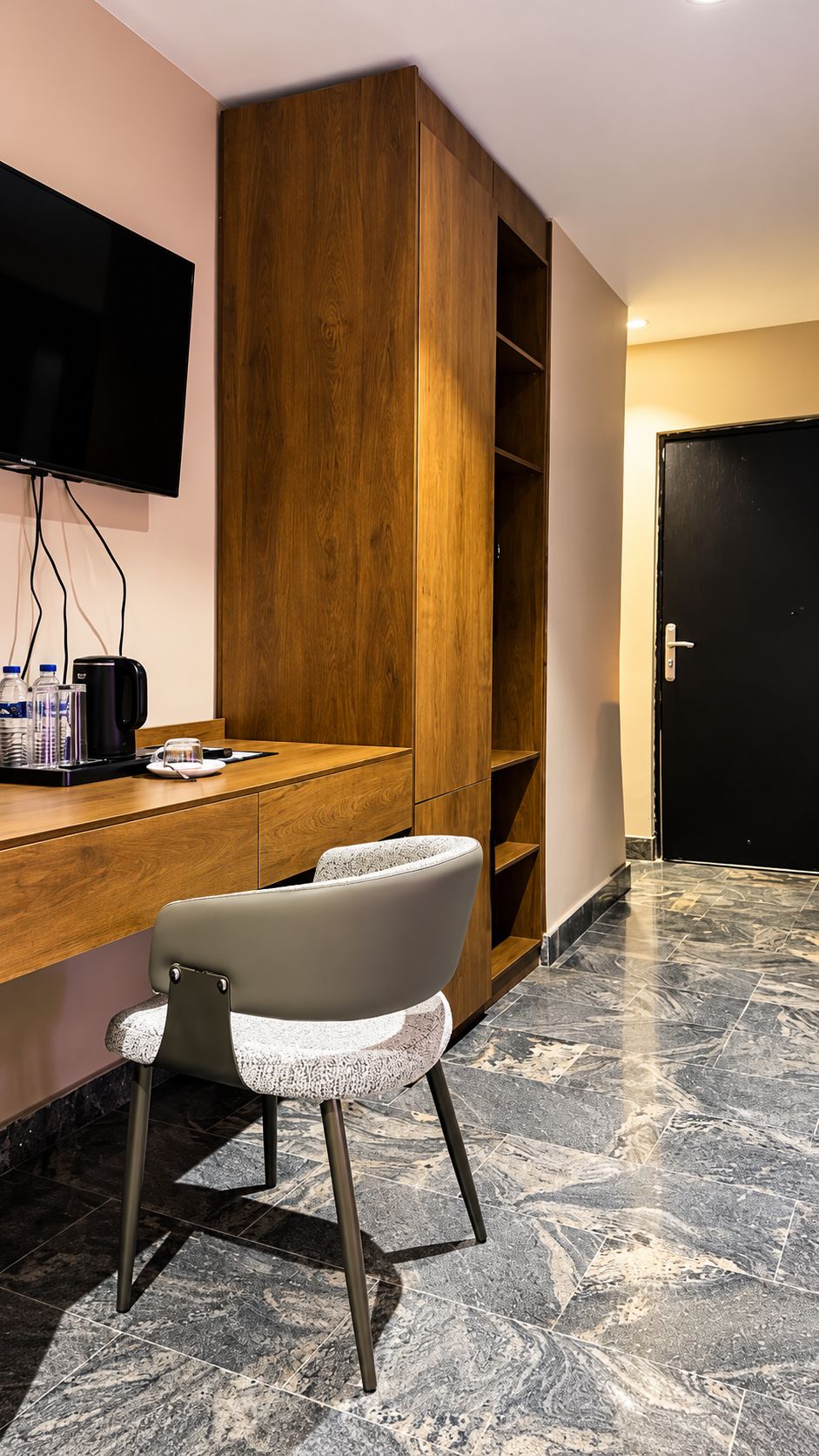 Habitación de hotel con escritorio de madera, televisor de pantalla plana montado en la pared, una silla moderna y piso con diseño de mármol.