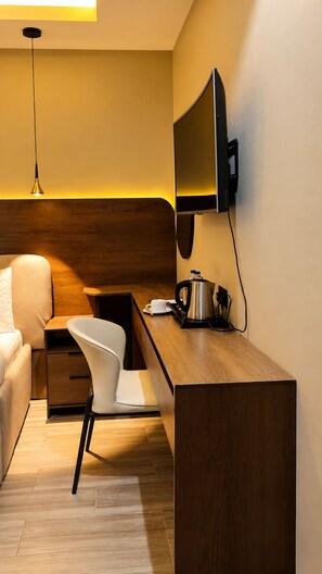 A modern hotel room with a wooden desk, a white chair, a bed with a beige headboard, and a flat-screen TV mounted on the wall. - Residency Hotel Lekki Lagos (Lekki)