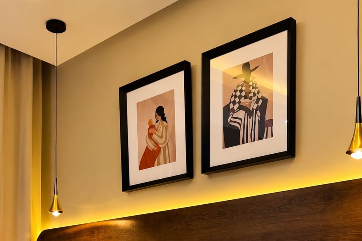 A hotel room with a bed, a headboard, two framed pictures on the wall, and a bedside table with a lamp.