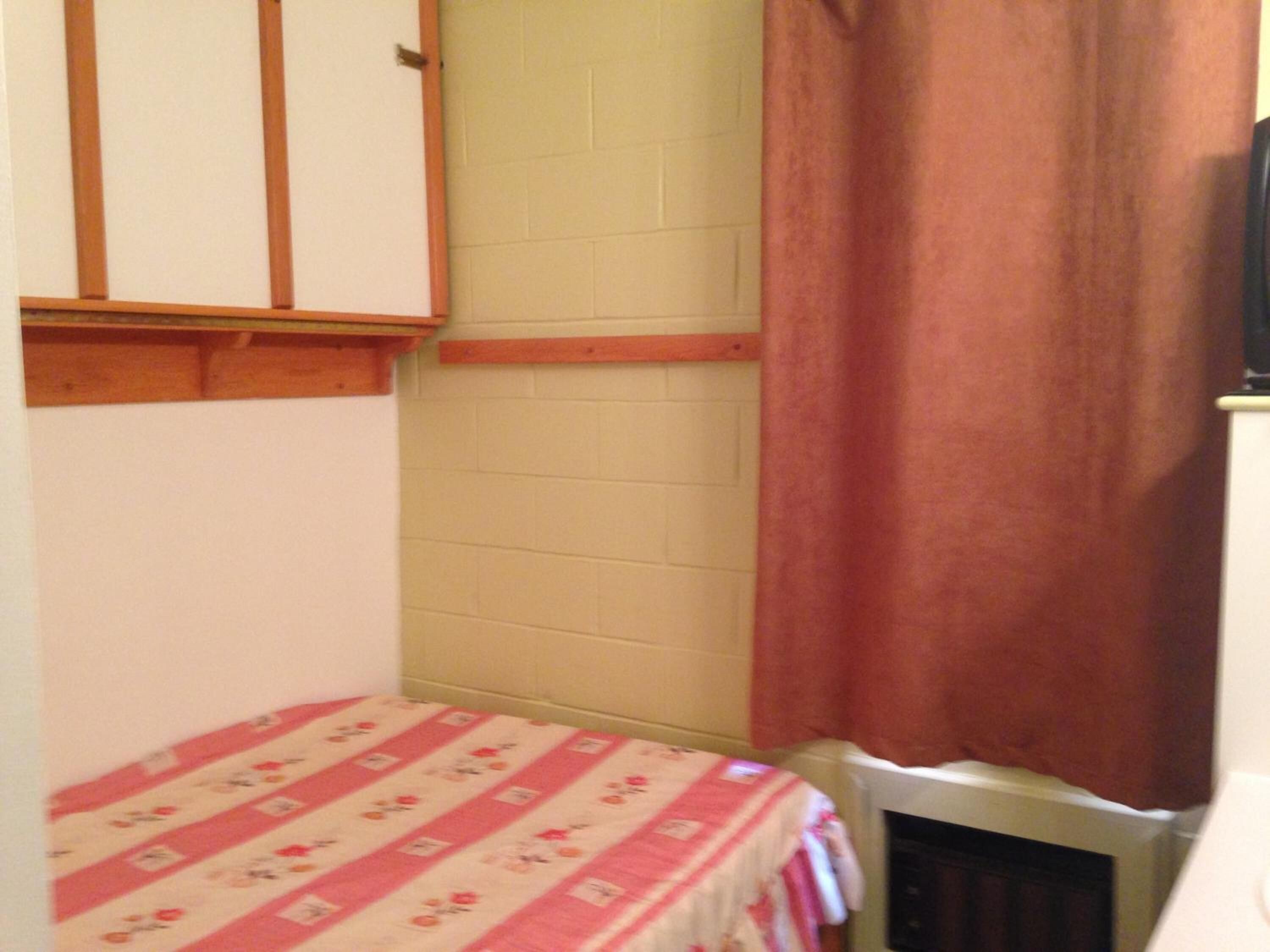 Quarto pequeno e estreito com uma cama simples coberta por um edredom estampado com flores. Uma prateleira de madeira está fixada na parede acima da cama, e um radiador branco é visível à direita.