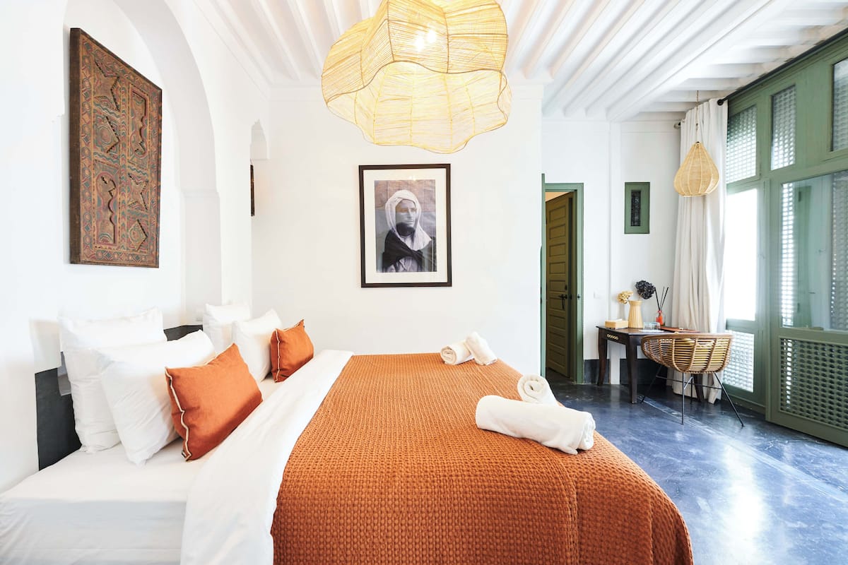 A hotel room with a large bed, a desk, and a chair. There is a window with curtains, a hanging light fixture, and a framed picture on the wall.