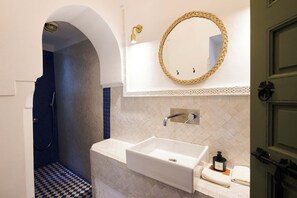 A bathroom with a white sink, a mirror with a woven frame, a shower with a blue wall, and a patterned floor. - Riad Dar Nakous (Marrakech)