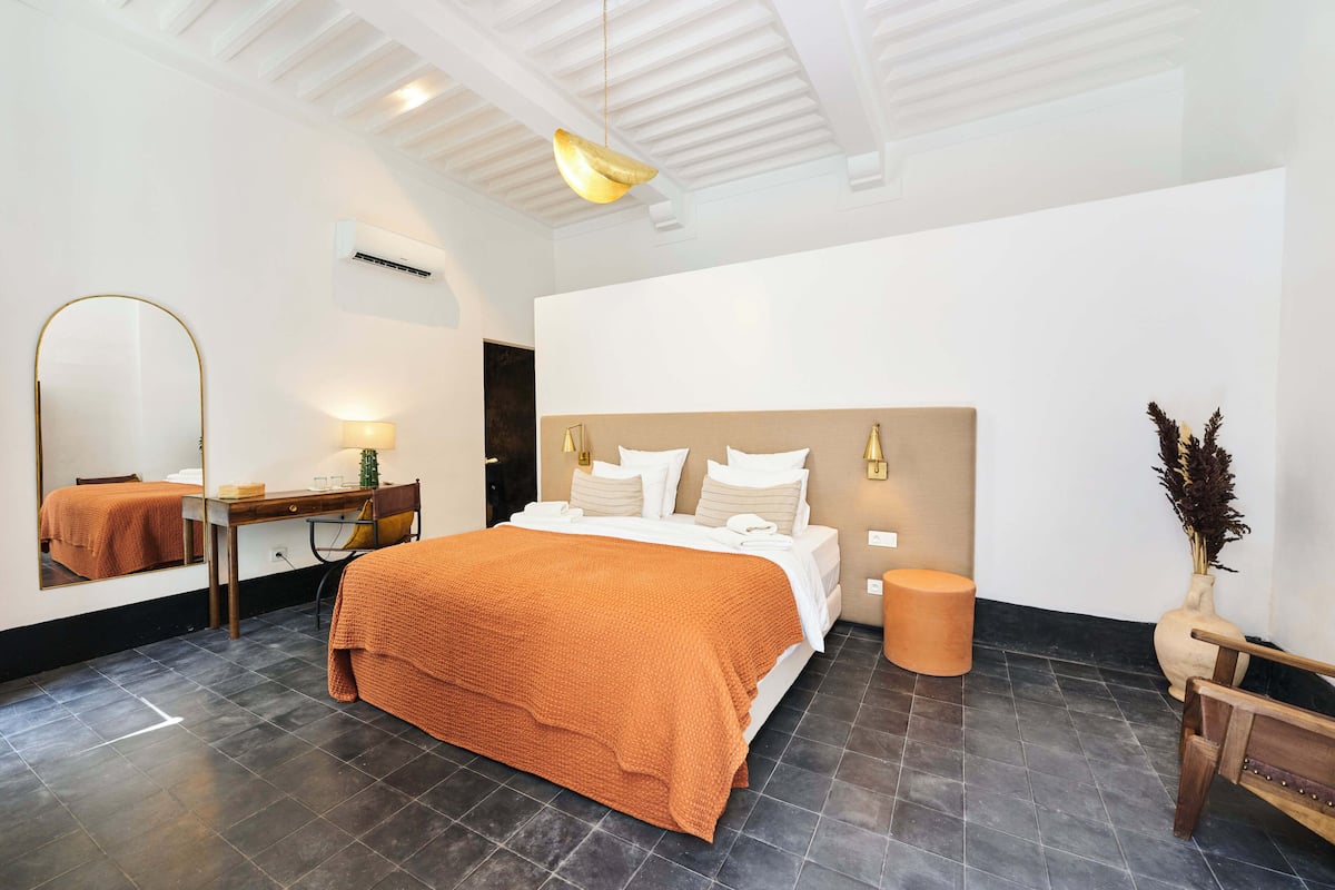 A modern hotel room with a large bed, a desk with a chair, a mirror, a bedside table, and a vase with dried plants.