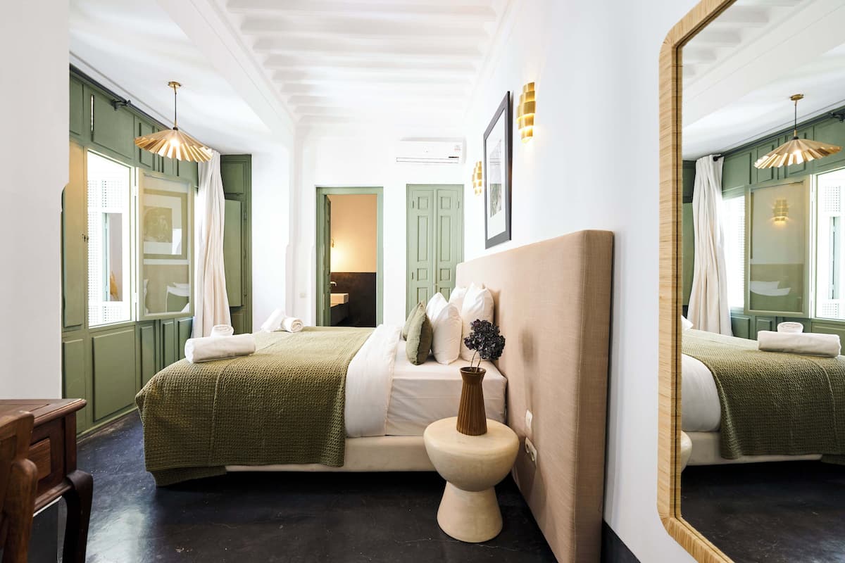 A hotel room with a large mirror, two beds with green bedding, a wooden nightstand, and a vase of flowers.