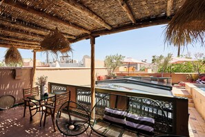 A terrace with a table and chairs, a thatched roof, and a view of buildings in the background. - Riad Dar Nakous (Marrakech)
