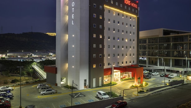 Un hotel moderno con un llamativo letrero de 'ibis HOTEL', varios pisos y un estacionamiento con varios coches.