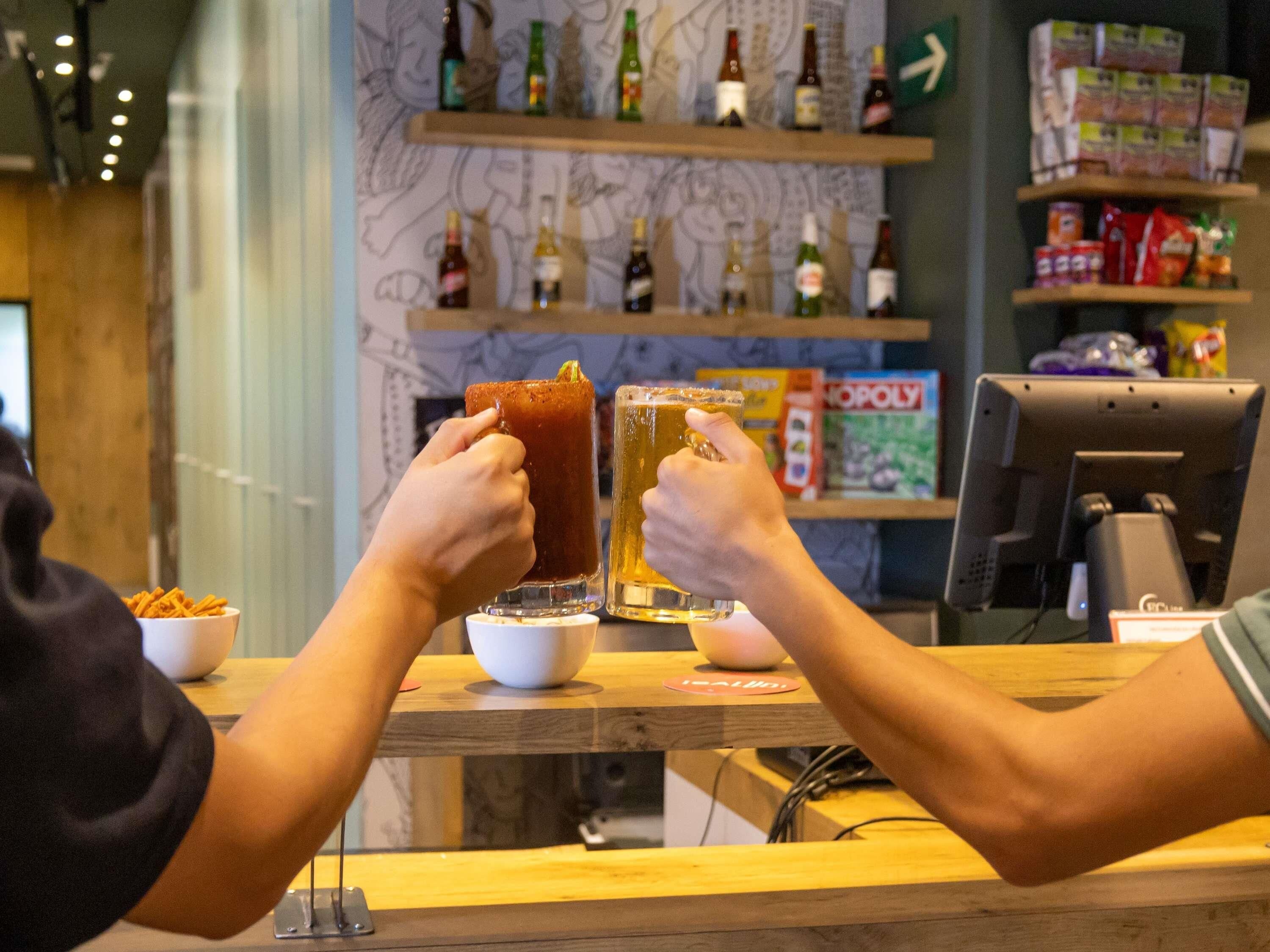 A bar area with two people holding drinks, a counter with a bowl of snacks, and shelves with bottles and snacks in the background.