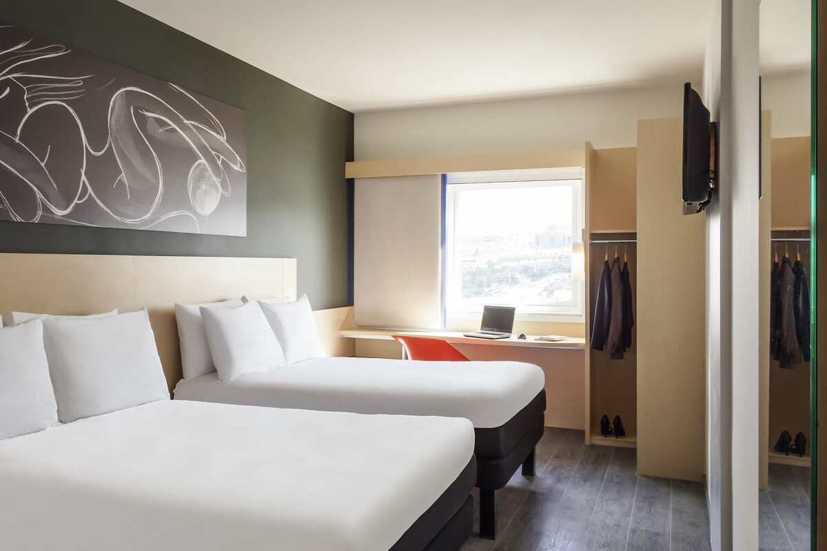 A hotel room with two beds, a desk with a laptop, a coat rack, and a window with a city view.