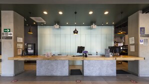 A reception desk with a modern design, featuring a light-colored wood top and a concrete base. There are two computer monitors, a printer, and a key box on the desk. The ceiling has recessed lighting and pendant lights. - ibis Queretaro (Querétaro)