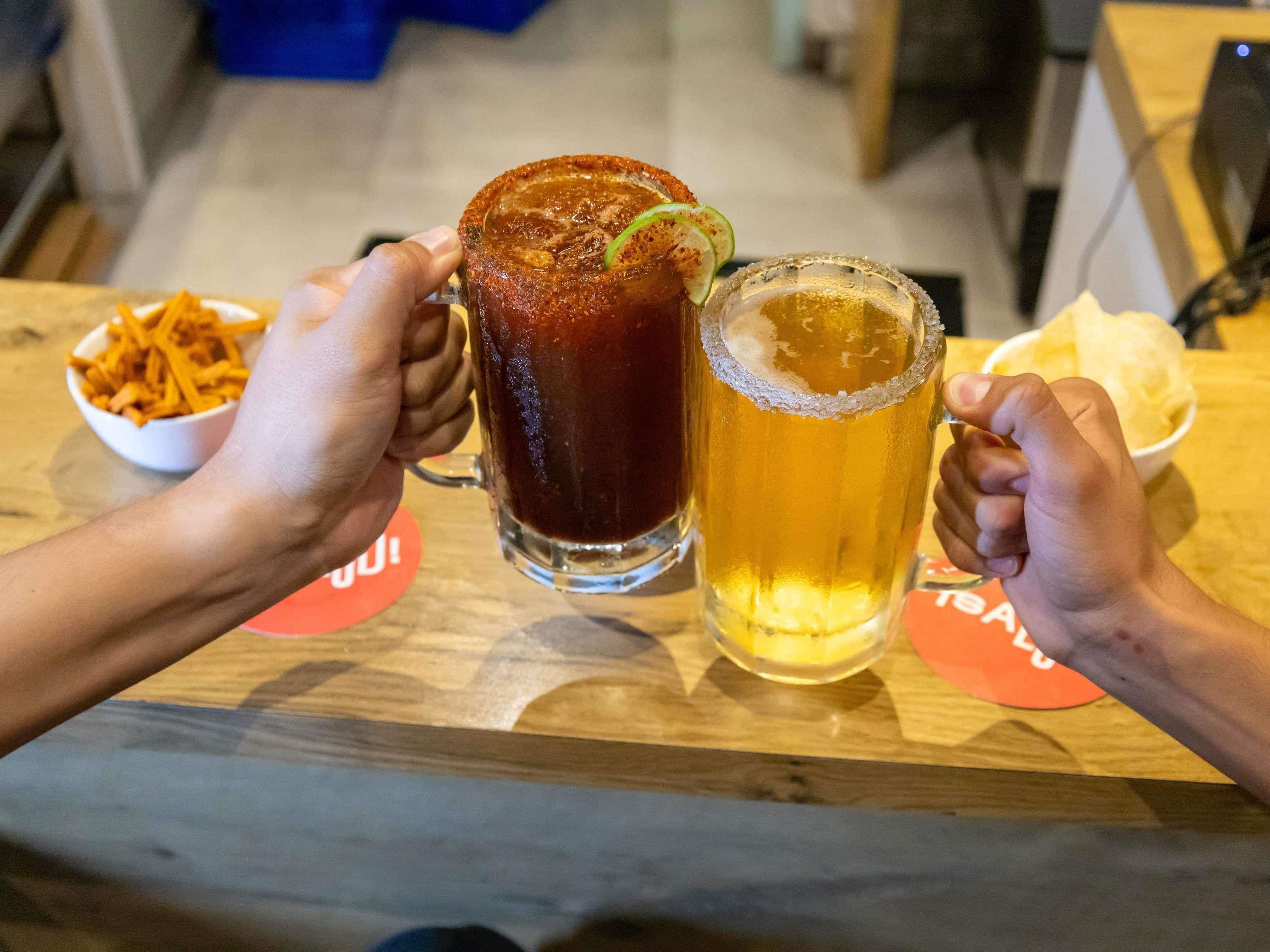 Dos manos sosteniendo un vaso alto y uno bajo de cerveza, con un tazón de aperitivos y una servilleta sobre la mesa.
