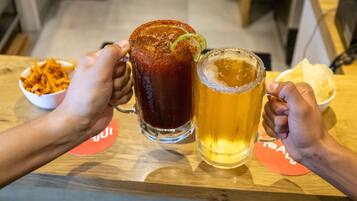 Dos manos sosteniendo un vaso alto y uno bajo de cerveza, con un tazón de aperitivos y una servilleta sobre la mesa.