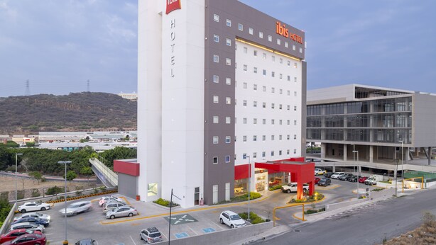 A modern hotel with a prominent "ibis HOTEL" sign, a parking lot with several cars, and a view of a hilly landscape in the background. - ibis Queretaro (Querétaro)